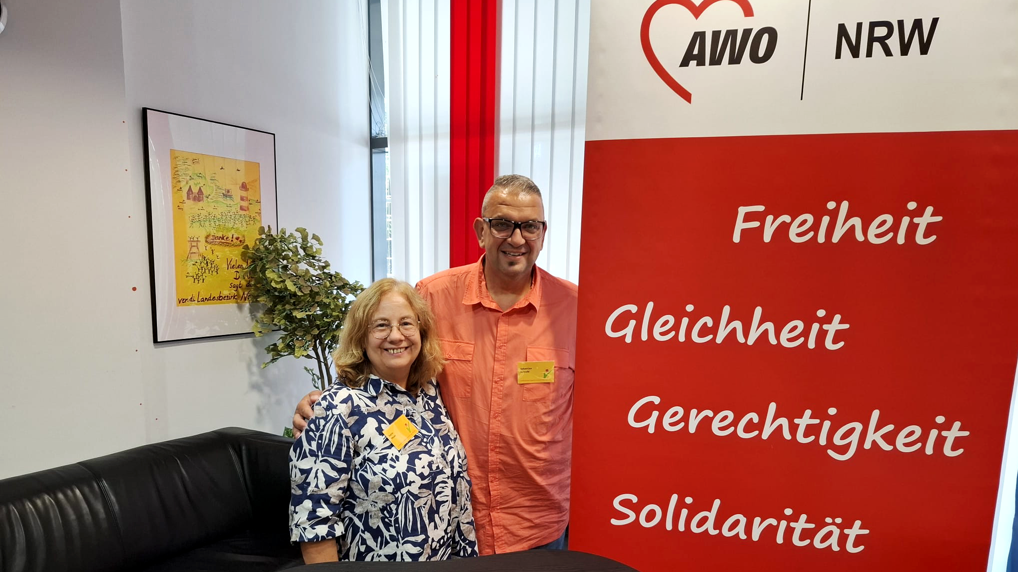 Auf dem Foto sind zwei freundlich lächelnde Personen zu sehen, die in einem Innenraum neben einem AWO-NRW-Roll-Up stehen. Die Frau trägt ein blau-weiß gemustertes Hemd, der Mann ein lachsfarbenes Hemd – beide tragen Namensschilder, was auf eine Veranstaltung oder ein Treffen hinweist. Das Roll-Up der AWO NRW (Arbeiterwohlfahrt Nordrhein-Westfalen) trägt in großer Schrift zentrale Werte der Organisation: Freiheit Gleichheit Gerechtigkeit Solidarität Toleranz Im Vordergrund befindet sich ein kleiner Infotresen mit mehreren AWO-Ausweisen oder Namenskarten und einem roten Kugelschreiber. Im Hintergrund ist ein Sofa zu sehen sowie ein gerahmtes gelbes Plakat mit Kinderzeichnungen und Schriftzügen, was auf ein kindgerechtes oder soziales Thema hinweist. Auch eine künstliche Pflanze ist dekorativ im Raum platziert. Insgesamt wirkt die Szene herzlich, offen und engagiert – ganz im Sinne der Werte der AWO. 