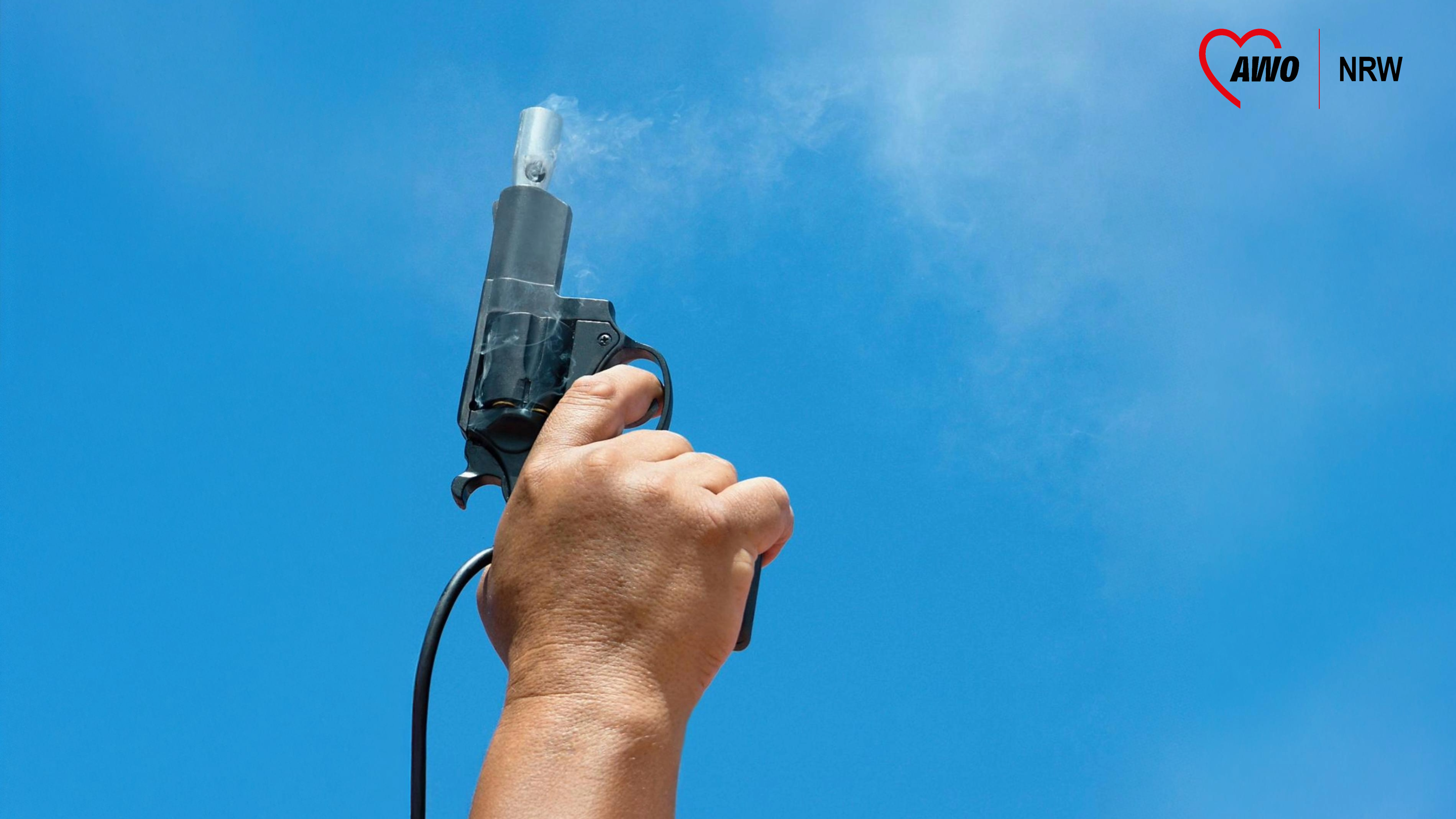 Auf dem Foto ist eine Hand zu sehen, die eine Startpistole in den Himmel hält. Die Pistole scheint gerade abgefeuert worden zu sein, da Rauch aus dem Lauf austritt. Der Hintergrund besteht aus einem klaren blauen Himmel. Außerdem ist oben rechts das Logo der AWO NRW zu sehen. 