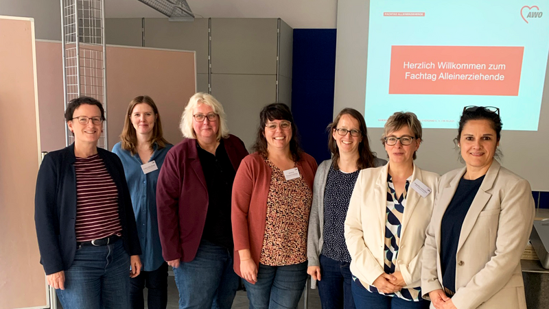 Auf dem Foto sind sieben Frauen in einem Innenraum zu sehen, die in einer Reihe stehen und in die Kamera schauen. Im Hintergrund gibt es eine Projektionsfläche oder einen Bildschirm, auf dem in deutscher Sprache in Rot und Weiß steht: Herzlich Willkommen zum Fachtag Alleinerziehende AWO (mit Logo) Die Frauen stehen vor der Leinwand. Der Raum hat helle Wände und einen hellen Boden. Es sind auch einige mobile Trennwände und Metallrahmenstrukturen im Raum erkennbar. Die Kleidung der Frauen ist eher leger und geschäftsmäßig, hauptsächlich bestehend aus Jeans und Oberteilen mit Blazern oder Strickjacken. Mehrere Personen tragen Namensschilder.