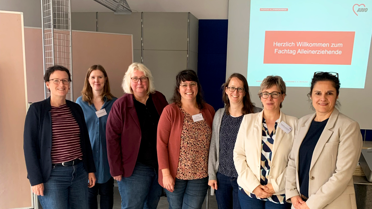Auf dem Foto sind sieben Frauen in einem Innenraum zu sehen, die in einer Reihe stehen und in die Kamera schauen. Im Hintergrund gibt es eine Projektionsfläche oder einen Bildschirm, auf dem in deutscher Sprache in Rot und Weiß steht: Herzlich Willkommen zum Fachtag Alleinerziehende AWO (mit Logo) Die Frauen stehen vor der Leinwand. Der Raum hat helle Wände und einen hellen Boden. Es sind auch einige mobile Trennwände und Metallrahmenstrukturen im Raum erkennbar. Die Kleidung der Frauen ist eher leger und geschäftsmäßig, hauptsächlich bestehend aus Jeans und Oberteilen mit Blazern oder Strickjacken. Mehrere Personen tragen Namensschilder.