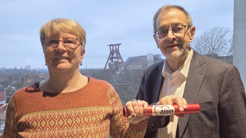 Auf dem Bild sind zwei Personen zu sehen, eine Frau und ein Mann, die ein rotes Objekt in Form einer Rolle oder eines Staffelstabs gemeinsam in die Kamera halten. Auf diesem roten Objekt ist ein weißes Logo mit einem Herz und dem Schriftzug "AWÖ" zu erkennen. Die Personen stehen vor einem großen Fenster. Im Hintergrund, außerhalb des Fensters, ist eine Industrieanlage oder ein Fördergerüst (ähnlich dem eines Bergwerks) zu sehen, was auf eine städtische oder industrielle Umgebung hindeutet. Die Umgebung wirkt wie eine Aussicht von einem oberen Stockwerk oder einem Balkon, und die Lichtverhältnisse deuten auf die Dämmerung oder einen bedeckten Tag hin.