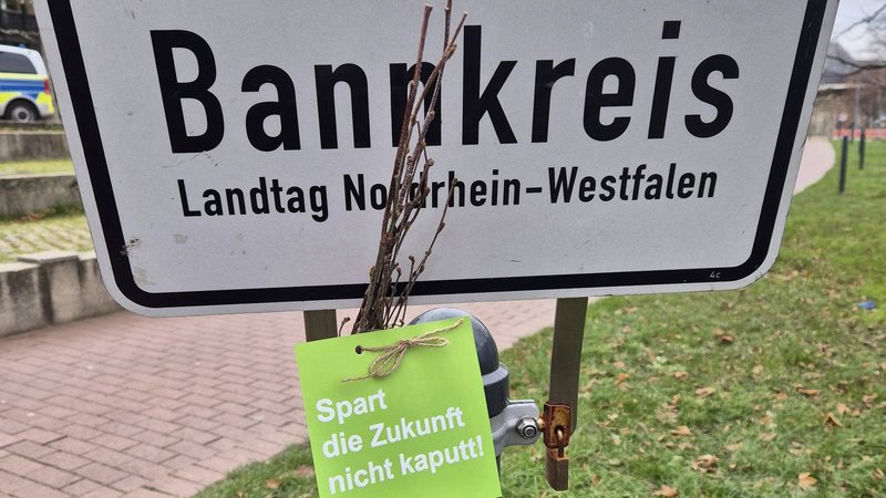 Das Foto zeigt ein Schild, mit den Worten "Bankkreis" und "Landtag Nordrhein-Westfalen". Vor dem Schild ist ein grüner Anhänger befestigt, auf dem steht: "Spart die Zukunft nicht kaputt!". Daneben ist ein Bündel kleiner Zweige angebracht.