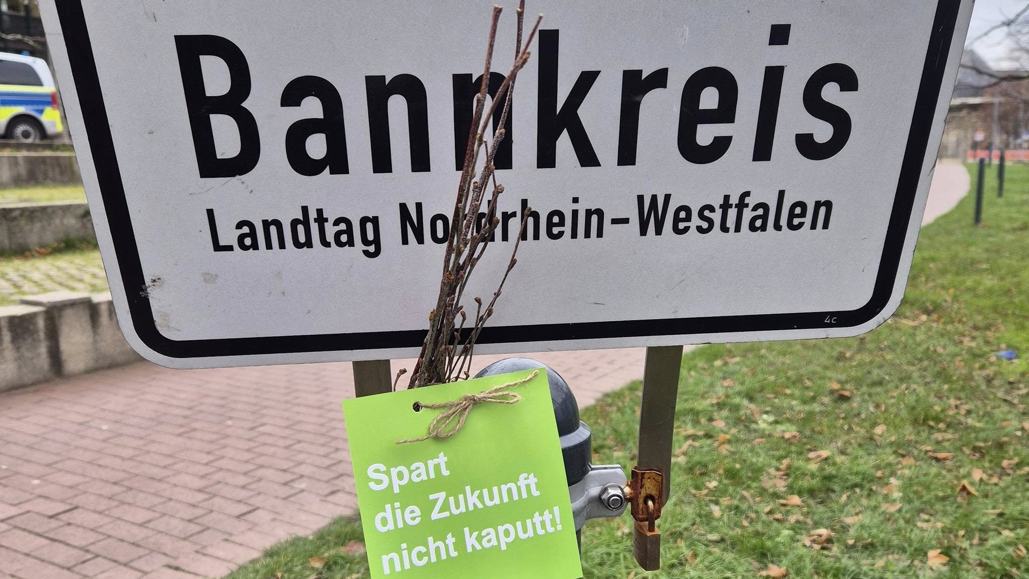 Das Foto zeigt ein Schild, mit den Worten "Bankkreis" und "Landtag Nordrhein-Westfalen". Vor dem Schild ist ein grüner Anhänger befestigt, auf dem steht: "Spart die Zukunft nicht kaputt!". Daneben ist ein Bündel kleiner Zweige angebracht.