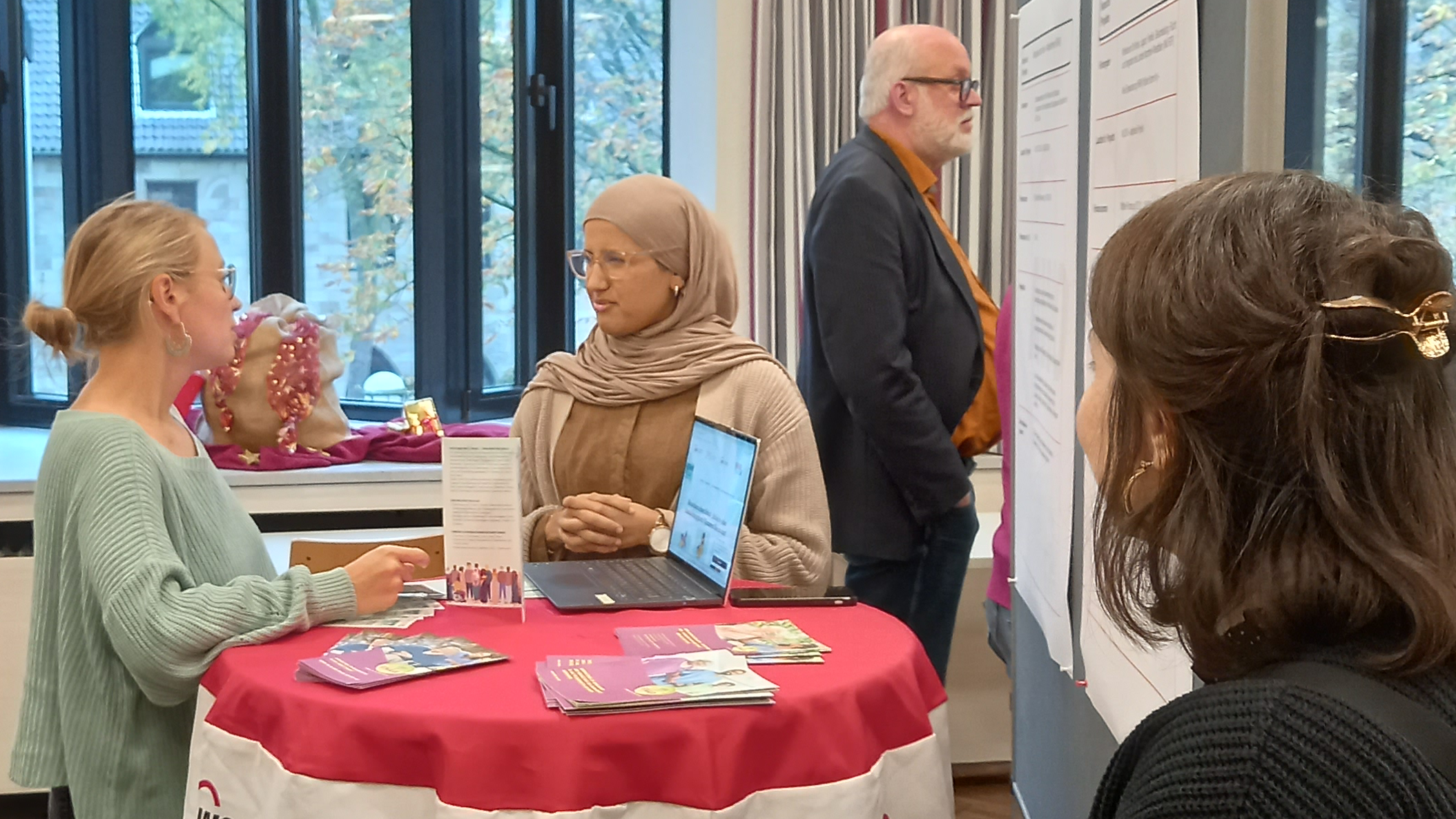 Das Foto zeigt eine Szene in einem Raum mit großen Fenstern im Hintergrund. Es sind mehrere Personen zu sehen, die an einem Tisch stehen, der mit einem roten Tuch bedeckt ist. Auf dem Tuch ist das Logo der Arbeiterwohlfahrt (AWO) zu erkennen. Auf dem Tisch liegen Broschüren und ein Laptop. Eine Person steht hinter dem Tisch und spricht mit einer anderen Person, die davor steht. Im Hintergrund schaut sich eine weitere Person eine Pinnwand mit Informationen an.