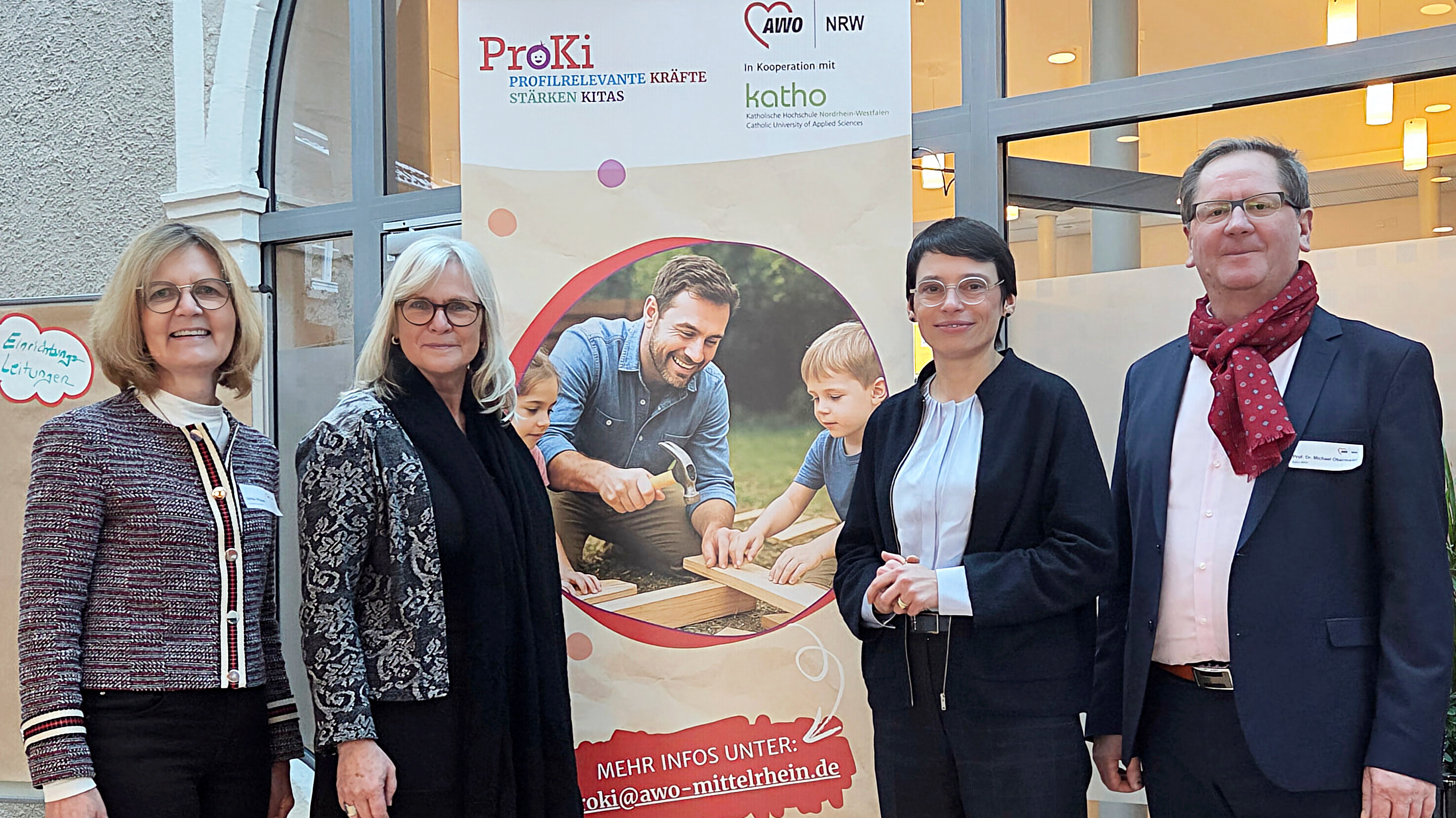 Auf dem Bild ist eine Gruppe von vier Personen (zwei Frauen, zwei Männer) vor einem großen Banner oder Plakat zu sehen. Die Personen stehen nebeneinander und blicken in die Kamera. Details zum Banner: Das Banner bewirbt ein Projekt mit dem Namen ProKi und trägt den Untertitel: "PROfilrelevante Kräfte im Kita stärken". Es zeigt die Kooperationspartner: AWO NRW (Arbeiterwohlfahrt Nordrhein-Westfalen) kath.o (Katholische Hochschule Nordrhein-Westfalen / Catholic University of Applied Sciences) In der Mitte des Banners ist dasselbe Motiv wie auf dem Banner im vorherigen Bild zu sehen: Ein lächelnder Mann, der zusammen mit zwei Kindern an einem Holzprojekt arbeitet. Am unteren Rand des Banners ist eine Kontaktinformation angegeben: MEHR INFOS UNTER: proki@awo-mittelrhein.de Die Szenerie findet wahrscheinlich in einem Foyer oder einem größeren Eingangsbereich statt, möglicherweise im Rahmen der Veranstaltung, die im vorherigen Bild gezeigt wurde.
