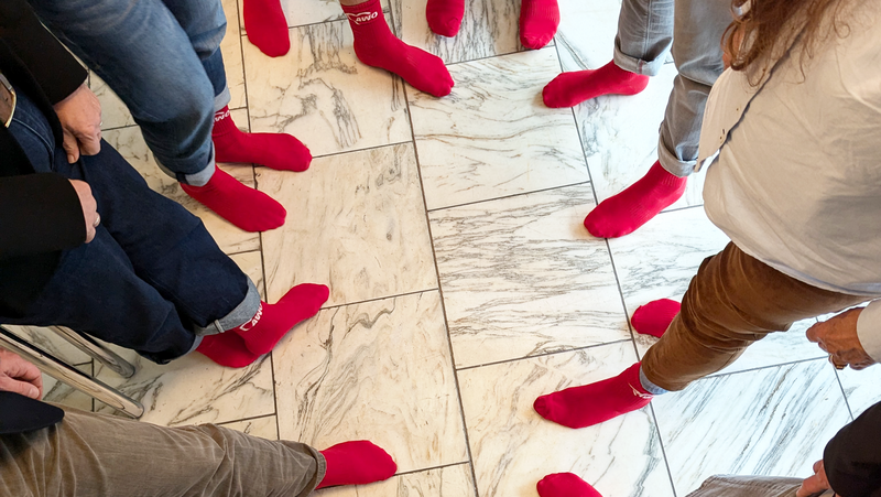 Das Foto zeigt eine Gruppe von Menschen, die in einem Kreis stehen. Sie tragen alle leuchtend rote Socken und sind von der Hüfte abwärts zu sehen. Der Boden besteht aus weiß-marmorierten Fliesen, was einen starken Kontrast zu den roten Socken bildet. 