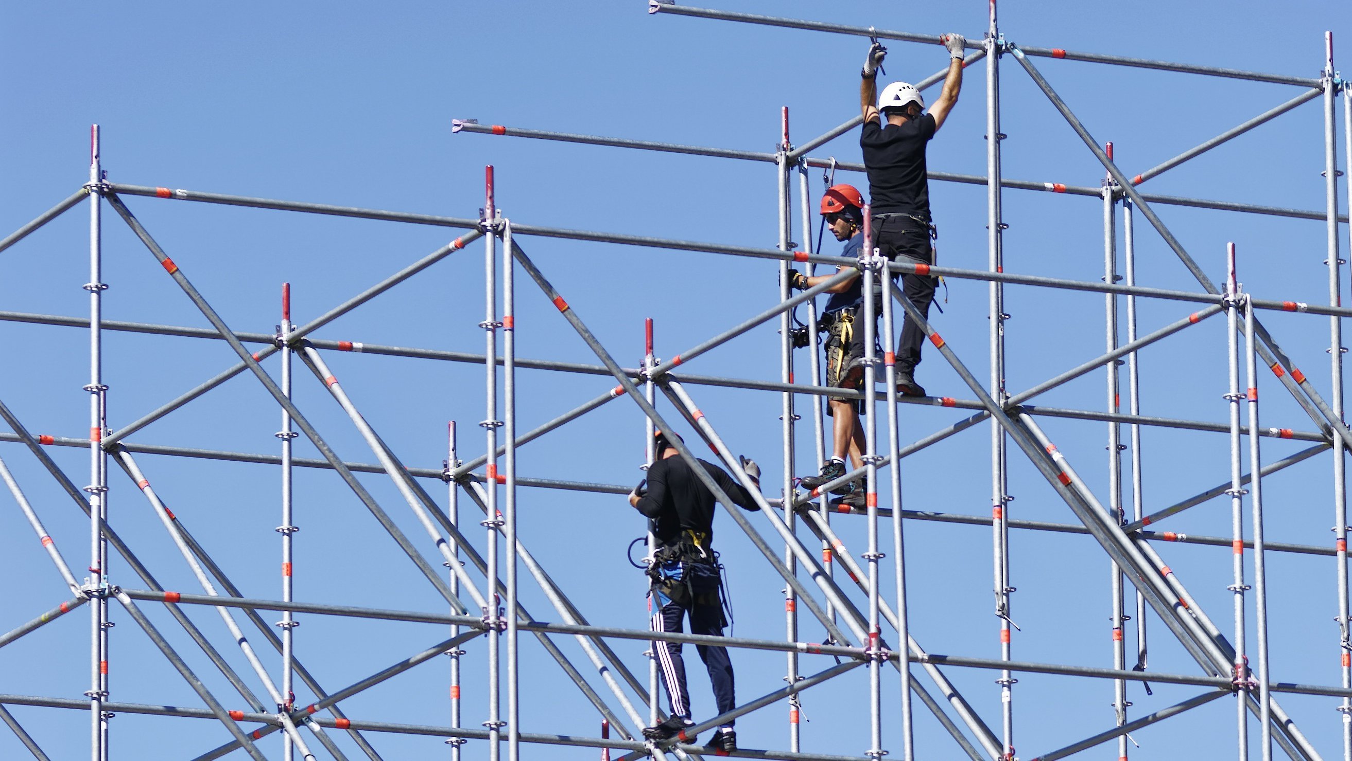 Auf dem Foto sind mehrere Bauarbeiter zu sehen, die an einem Gerüst arbeiten. Sie montieren oder sichern Metallstangen und sind mit Sicherheitsausrüstung ausgestattet, darunter Helme, Gurte und Werkzeuge. Das Gerüst ist hoch und komplex aufgebaut. Im Hintergrund sind ein Gebäude und Bäume zu erkennen, und der Himmel ist klar und blau.