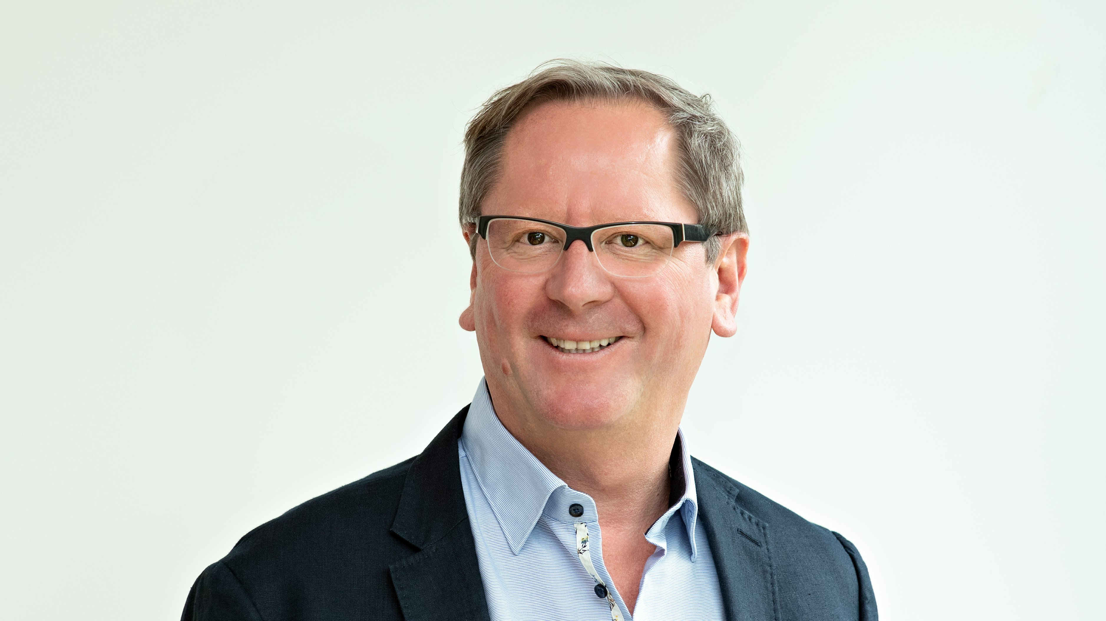 Auf dem Bild ist ein Mann zu sehen. Er trägt eine Brille, ein hellblaues Hemd und ein dunkelblaues Sakko. Er wirkt freundlich und lächelt in die Kamera. Der Hintergrund ist schlicht und hell, sodass der Fokus ganz auf der Person liegt.