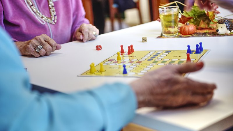 Auf dem Foto sind mehrere ältere Personen zu sehen, die gemeinsam ein Brettspiel spielen. Es handelt sich um das Spiel „Mensch ärgere dich nicht“. Auf dem Tisch liegt das Spielbrett mit bunten Spielfiguren (gelb, blau, rot) und zwei Würfeln. Die Szene spielt sich vermutlich in einem Gemeinschaftsraum ab – vielleicht in einem Seniorenheim oder einer Betreuungseinrichtung. Im Hintergrund steht ein Glas mit einem Getränk und herbstlicher Dekoration (z. B. kleine Kürbisse und ein Igel). Die Kleidung und der Schmuck der Personen sowie die liebevolle Tischdekoration lassen eine warme, gepflegte Atmosphäre erkennen. Das Bild vermittelt Gemeinschaft, Aktivität und Lebensfreude im Alter.