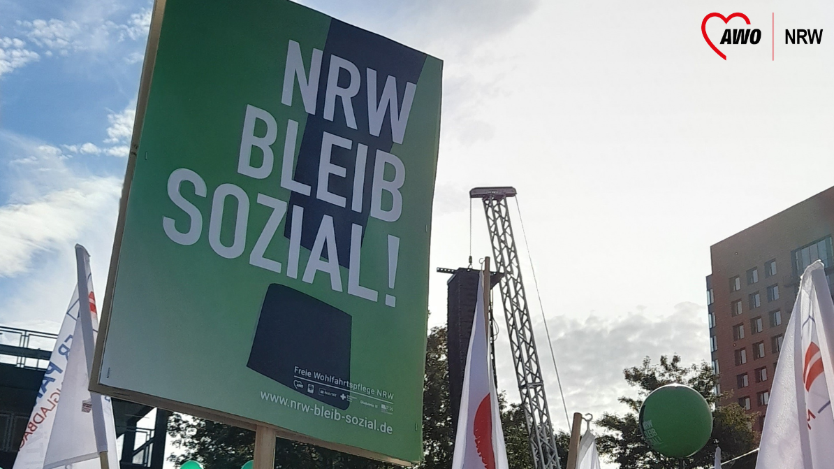 Das Bild zeigt ein Plakat mit der Aufschrift „NRW BLEIB SOZIAL!“ in großen weißen Buchstaben auf grünem Hintergrund. Unten auf dem Plakat sind die Logos verschiedener Wohlfahrtsverbände zu erkennen, die zur Freien Wohlfahrtspflege in Nordrhein-Westfalen (NRW) gehören, wie das der Arbeiterwohlfahrt (AWO). Die URL „nrw-bleib-sozial.de“ ist ebenfalls unten auf dem Plakat zu sehen. Im Hintergrund sind weitere Fahnen und Menschen zu sehen, was auf eine Veranstaltung oder Demonstration hindeutet.