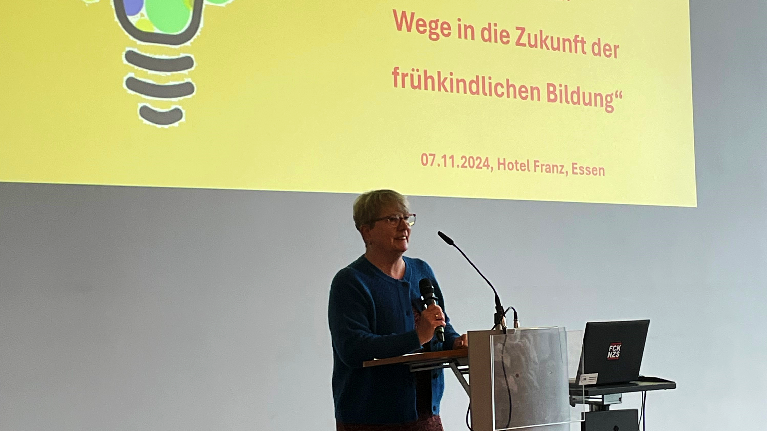 Auf dem Foto ist Britta Altenkamp zu sehen, die an einem Rednerpult steht und in ein Mikrofon spricht. Hinter ihr ist eine Leinwand mit einer Präsentation projiziert, auf der der Titel „Vision Kita: Wege in die Zukunft der frühkindlichen Bildung“ und das Datum „07.11.2024, Hotel Franz, Essen“ zu lesen sind. 