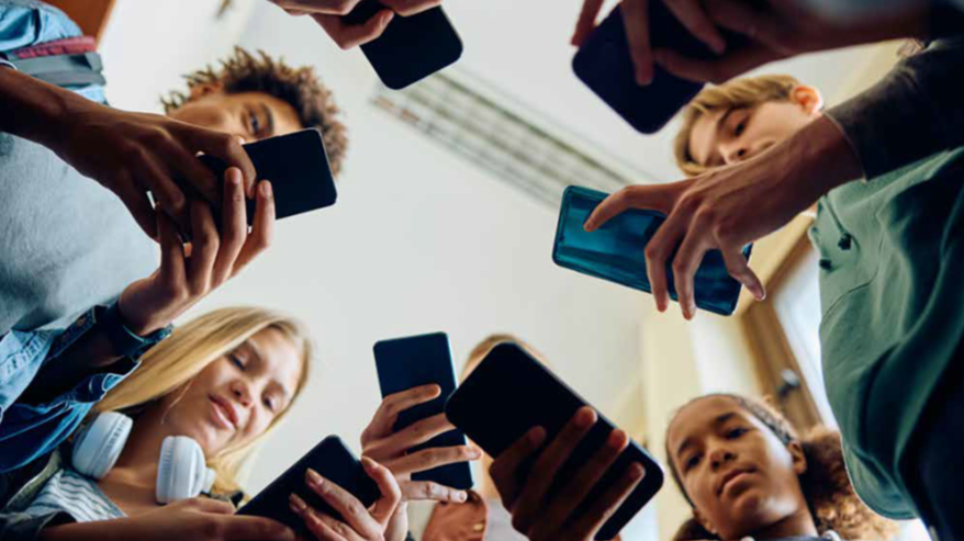Auf dem Bild ist eine Gruppe von etwa sieben Jugendlichen zu sehen, die in einem Kreis stehen und von unten fotografiert wurden. Alle Personen schauen nach unten auf ihr Smartphone, das sie in den Händen halten. Die Bildschirme der Telefone sind beleuchtet. Die Szene vermittelt den Eindruck, dass die Gruppe stark auf ihre mobilen Geräte konzentriert ist und miteinander durch die Telefone interagiert oder sie gemeinsam benutzt. Im Hintergrund sind verschwommen eine Decke und eventuell ein Fenster oder eine Lichtquelle zu erkennen.