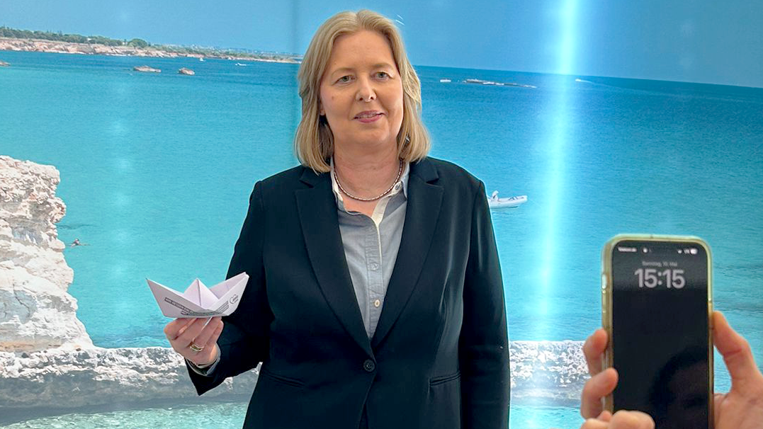 Auf dem Foto ist eine Frau in einem dunklen Blazer und einer hellgrauen Bluse zu sehen, die ein gefaltetes Papierboot in ihrer linken Hand hält. Sie steht vor einem Hintergrund, der eine Küstenlandschaft mit blauem Meer, hellem Sand und Felsen zeigt. Rechts im Bild ist ein Teil einer Person zu sehen, die ein Smartphone in beiden Händen hält und die Frau zu fotografieren scheint. Auf dem Display des Smartphones ist die Uhrzeit 15:15 zu erkennen. Links im Bild ist ein runder Tisch mit einigen Papierdokumenten darauf angeschnitten.