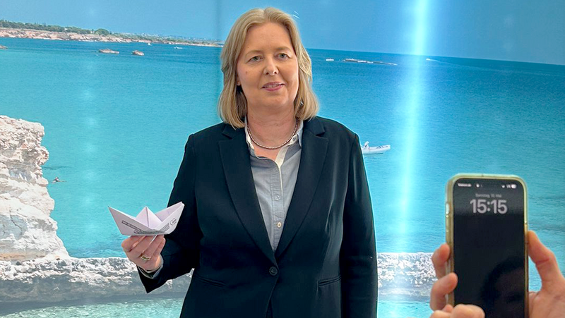 Auf dem Foto ist eine Frau in einem dunklen Blazer und einer hellgrauen Bluse zu sehen, die ein gefaltetes Papierboot in ihrer linken Hand hält. Sie steht vor einem Hintergrund, der eine Küstenlandschaft mit blauem Meer, hellem Sand und Felsen zeigt. Rechts im Bild ist ein Teil einer Person zu sehen, die ein Smartphone in beiden Händen hält und die Frau zu fotografieren scheint. Auf dem Display des Smartphones ist die Uhrzeit 15:15 zu erkennen. Links im Bild ist ein runder Tisch mit einigen Papierdokumenten darauf angeschnitten.