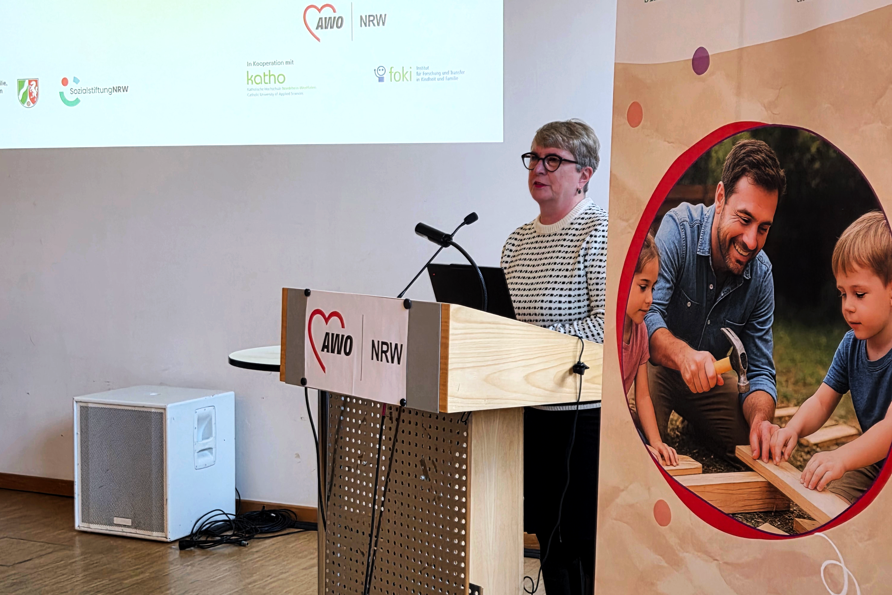 Auf dem Bild ist eine Veranstaltung oder Präsentation zu sehen. Hier sind die Details: Vordergrund: Eine Frau steht an einem Rednerpult und spricht. Sie trägt eine gestreifte Bluse und eine Brille. Rednerpult: Das Pult ist aus hellem Holz und trägt das Logo der AWO NRW (Arbeiterwohlfahrt Nordrhein-Westfalen), zusammen mit einem roten Herz-Symbol. Hintergrund (Leinwand/Projektion): Über der Frau ist eine Projektionsfläche oder eine Tafel mit weiteren Logos zu sehen, darunter: Sozialstiftung NRW AWO NRW Katho (Katholische Hochschule Nordrhein-Westfalen) foki (vermutlich ein Institut/Projekt) Rechte Seite: Ein großes Plakat oder Banner mit einem runden Ausschnitt zeigt einen lächelnden Mann, der zusammen mit zwei Kindern an einem Holzprojekt arbeitet (er hält einen Hammer). Linke Seite (Boden): Ein weißer Lautsprecher oder eine Box steht auf dem Holzboden.