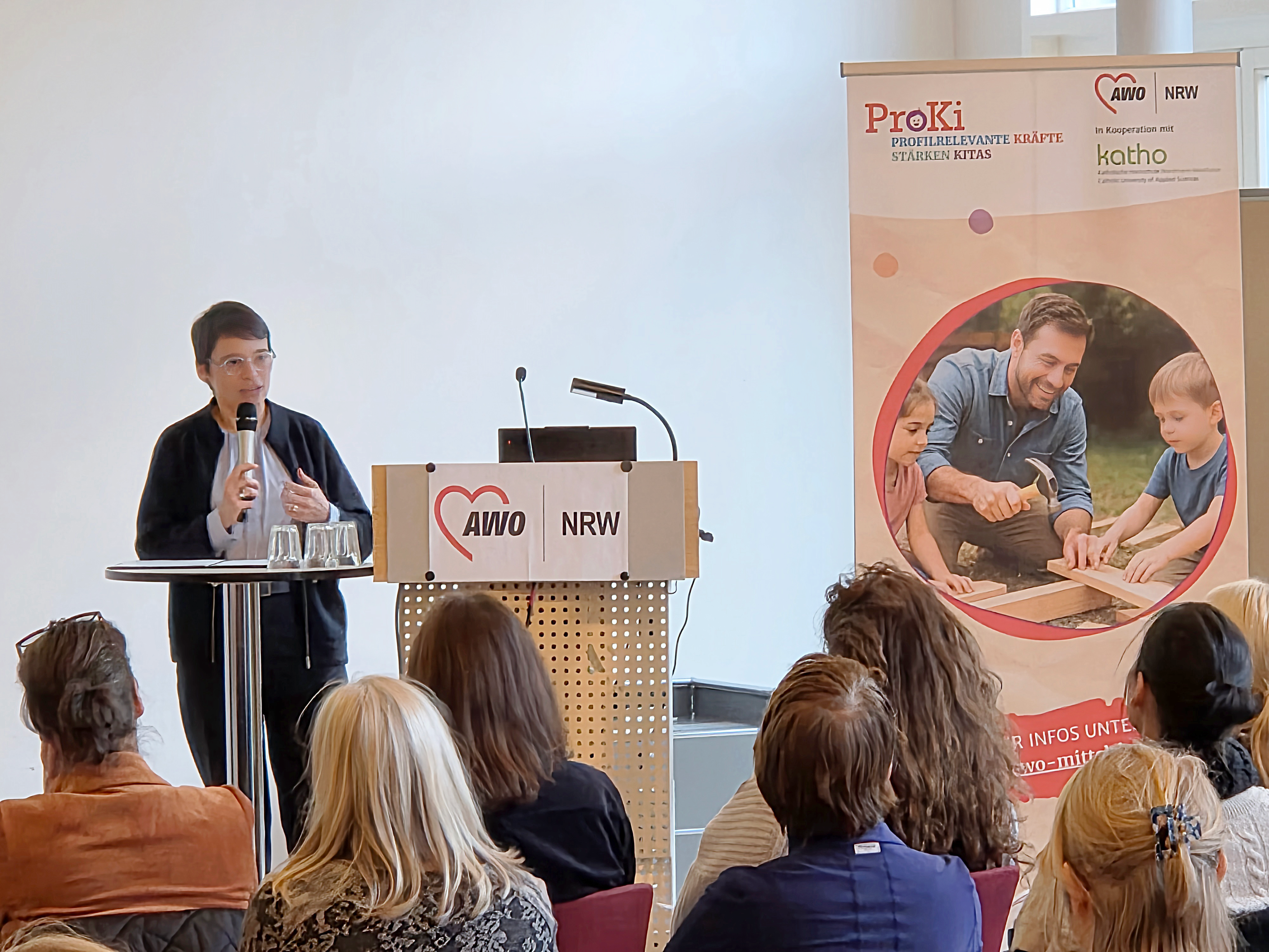 Auf dem Bild ist eine Präsentation oder ein Vortrag im Rahmen einer Veranstaltung zu sehen. Hier sind die Details: Vortragende: Eine Frau steht links im Bild an einem kleinen runden Tisch und hält ein Mikrofon. Sie scheint zu einem Publikum zu sprechen. Sie trägt eine helle Bluse unter einem dunklen Oberteil. Rednerpult: Hinter ihr steht ein größeres Rednerpult mit dem Logo der AWO NRW (Arbeiterwohlfahrt Nordrhein-Westfalen). Publikum: Im Vordergrund und in der Mitte des Bildes sind die Rücken des Publikums zu sehen, das dem Vortrag folgt. Banner/Plakat: Rechts steht ein großes Banner, das für das Projekt "ProKi – PROfilrelevante Kräfte im Kita stärken" wirbt. Die Kooperationspartner AWO NRW und kath.o (Katholische Hochschule NRW) sind darauf genannt. Das Motiv in der Mitte zeigt einen Mann, der mit zwei Kindern an einem Holzprojekt arbeitet. Die Szene zeigt offensichtlich einen Moment während der Veranstaltung, die durch die Bilder 1 und 2 dokumentiert wurde.