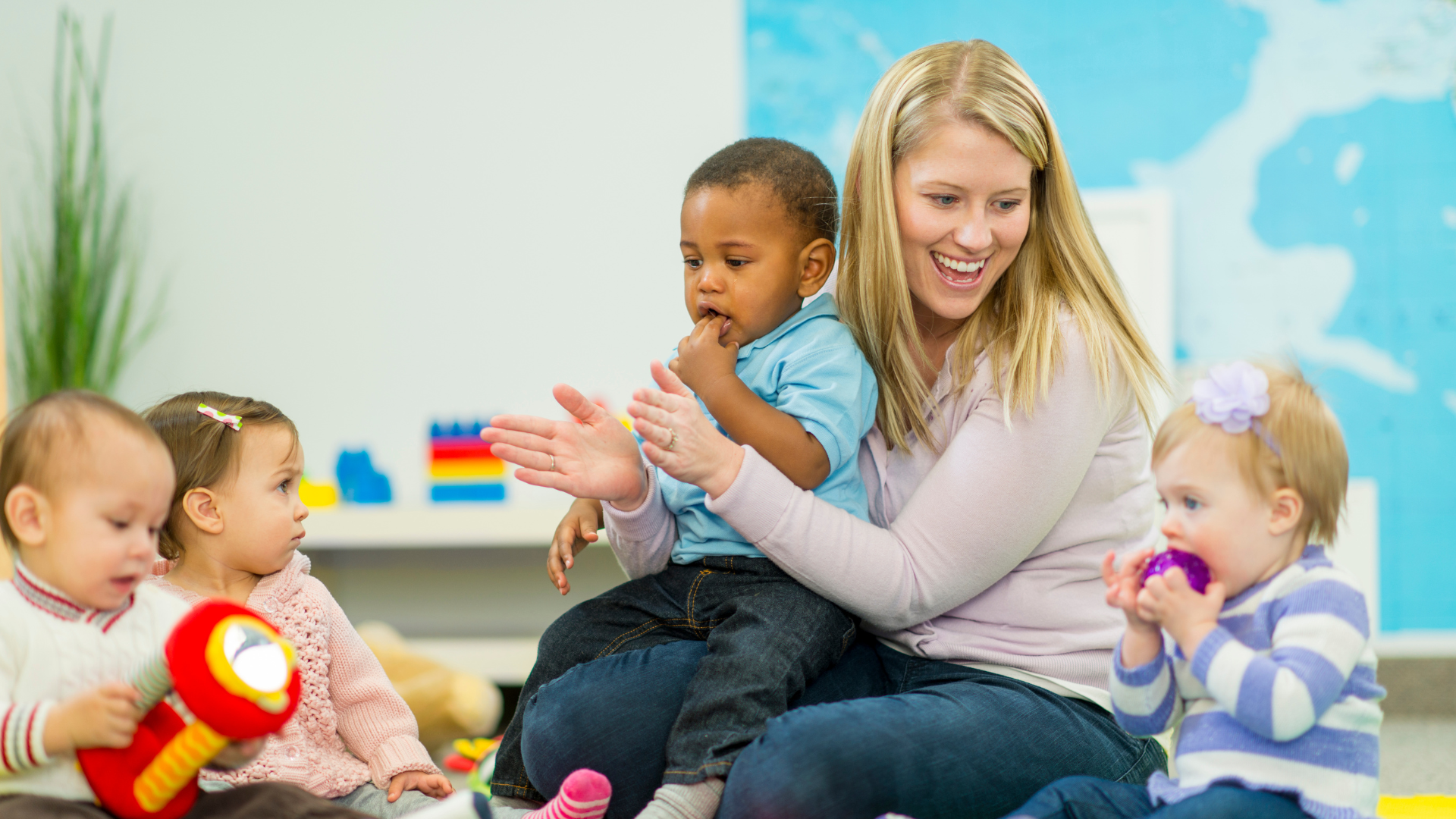  Auf dem Foto sind eine lächelnde blonde Frau und vier kleine Kinder zu sehen, die an einer Aktivität teilnehmen, möglicherweise in einer Kinderbetreuungseinrichtung oder einem Spielkreis. Die Frau hält ein Kleinkind auf ihrem Schoß, das seinen Finger im Mund hat. Drei weitere Kleinkinder sitzen auf dem Boden vor ihr. Eines der Kinder auf dem Boden hat ein rotes Spielzeug in der Hand, während ein anderes Kind ein lila Spielzeug zum Mund führt. Im Hintergrund ist eine große Karte oder eine ähnliche Darstellung zu sehen. Die allgemeine Atmosphäre des Fotos ist fröhlich und engagiert.